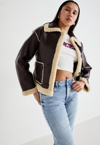 Bruine neplederen jas met beige schapenvachtvoering en -afwerking; heeft een kraag en twee voorkezen; gestyled met lichtblauwe high-waisted jeans.