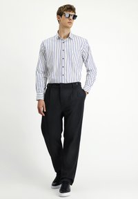Camicia bianca con righe blu, taschino frontale, abbottonata. Pantaloni neri plissettati, tasche laterali, abbinati a sneaker nere. Occhiali da sole indossati.