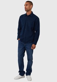 Marineblauwe longsleeve shirt met twee borstzakken, knoopsluiting en een relaxte pasvorm, gecombineerd met blauwe jeans en witte sneakers.