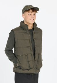 Olivgrüne, gesteppte Pufferweste mit hohem Kragen, zwei Reißverschlusstaschen und einem schwarzen Etikett. Über einem passenden Langarmshirt getragen.