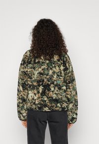 Kurtka z polaru w zielony, czarny i beżowy wzór moro. Posiada wysoki kołnierz i długie rękawy. Teksturowana tkanina o luźnym kroju.