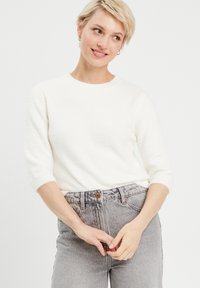 Pull en tricot blanc et duveteux avec des manches trois-quarts, un col rond et une texture lisse, associé à un jean taille haute gris clair.