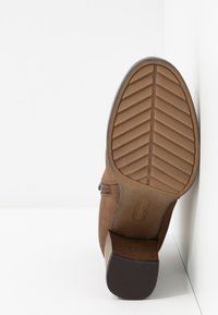 Botte cheville en daim marron avec semelle en caoutchouc montrant un motif de bande de roulement en forme de chevron et un talon empilé sur un fond blanc.