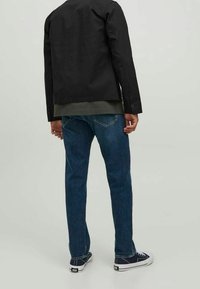 Svart jacka med en slät textur, grön skjorta under, blå denimjeans och marinblå sneakers. Synlig sömnad och knappar på jackan.