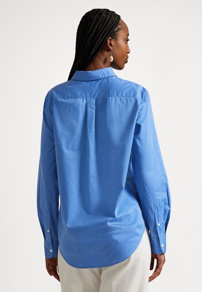 Hosszú ujjú kék inget és fehér nadrágot viselő, fonott hajú nő, háttal egy egyszínű fehér háttér előtt.