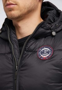 Chaqueta negra acolchada con cierre de cremallera, capucha con cordón y un parche bordado circular con el texto "QUIERO IRME."