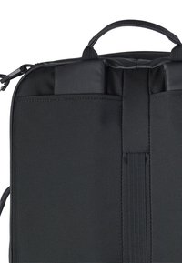 Svart texturerad tygryggsäck med vadderade remmar, handtag på toppen och vertikalt remsläpp mot en vit bakgrund.