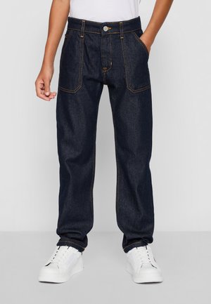 Personne portant un jean en denim bleu foncé avec poches plaquées à l'avant et des baskets blanches, debout devant un fond uni.