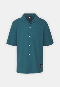 Camicia a maniche corte con bottoni in tessuto testurizzato di colore teal, caratterizzata da una singola tasca sul petto e bottoni bianchi, con etichetta del logo sull'orlo.