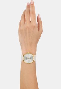 Runde Uhr mit silbernem Ziffernblatt, goldenen Akzenten und beigem Armband. Verfügt über klare Stundenmarkierungen und ein minimalistisches Design. Getragen an einer Hand.