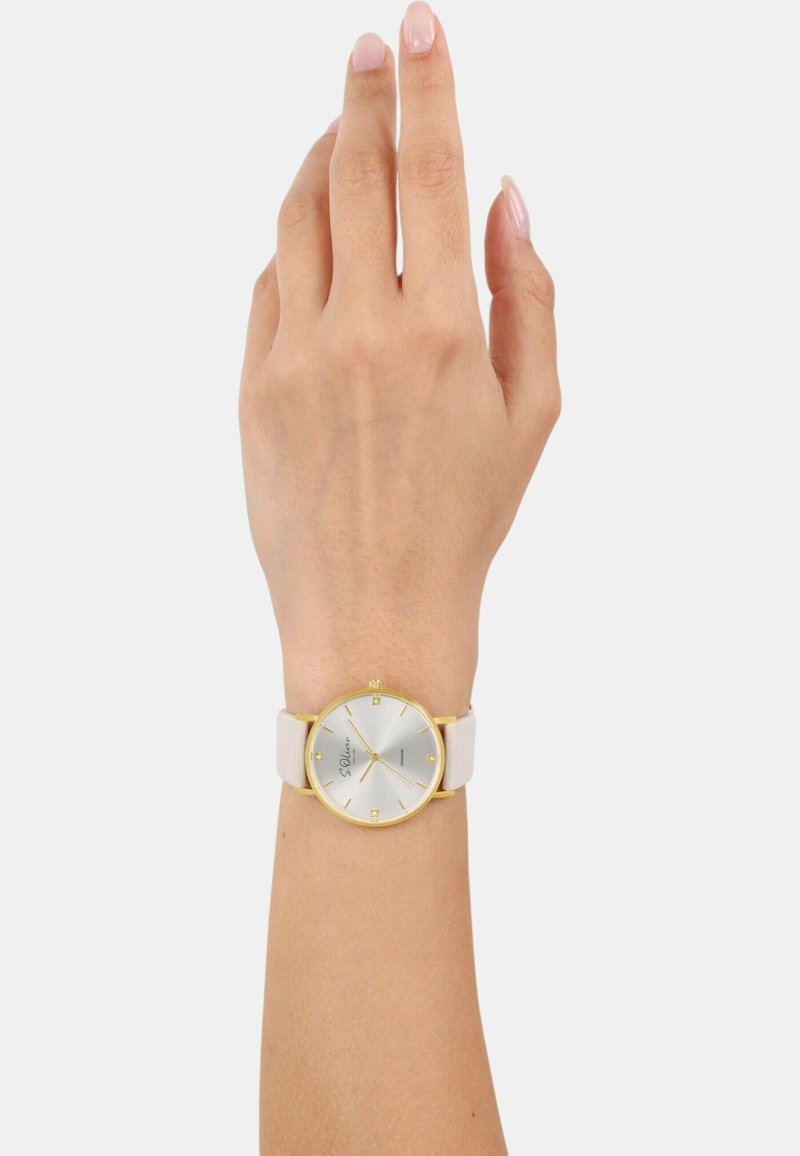 Runde Uhr mit silbernem Ziffernblatt, goldenen Akzenten und beigem Armband. Verfügt über klare Stundenmarkierungen und ein minimalistisches Design. Getragen an einer Hand.