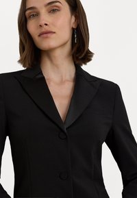 Juodas švarkas su satino atlapu, vienu mygtuko užsegimu, dviem priekinėmis kišenėmis ir struktūruotomis petukais. Lygus audinys, pritaikytas prie kūno.