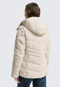 Chaqueta acolchada beige con capucha, que presenta un diseño acolchado y costuras diagonales. Fabricada con una tela suave y lisa, tiene un corte holgado.