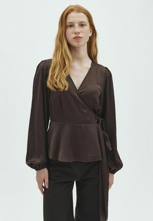 Jeune femme aux longs cheveux roux portant un chemisier cache-cœur marron foncé à manches longues et un pantalon noir sur un fond clair uni.