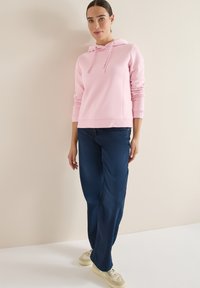 Felpa rosa chiaro con coulisse, polsini a costine, abbinata a jeans a gamba larga blu scuro e sneakers di colore chiaro. Tessuto dalla texture morbida.