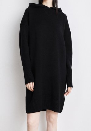 Woman wearing a loose black knitted hooded dress with long sleeves and ribbed cuffs and hem, standing against a plain light wall.