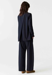 Ensemble costume rayé bleu marine composé d'une longue veste avec une fente au dos, associée à un pantalon à jambes larges. Le matériau semble lisse et ajusté.