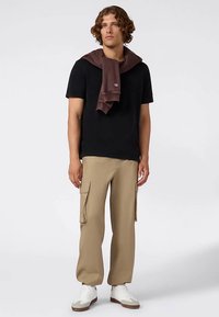 Camiseta negra de manga corta, suéter marrón colgado sobre los hombros, pantalones cargo color beige, zapatillas blancas con suelas marrones, postura de pie contra un fondo liso.