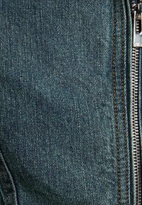 Tessuto denim blu con trama verticale visibile, caratterizzato da una cerniera argentata a destra e cuciture arancioni a contrasto lungo le cuciture.