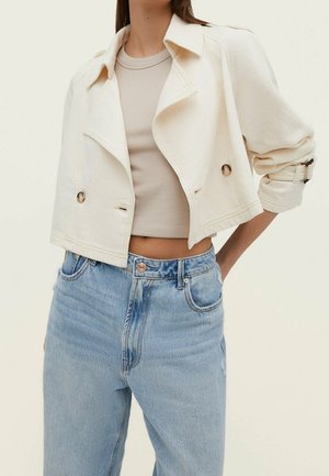 Femme portant une veste courte crème à double boutonnage, un crop top côtelé beige et un jean taille haute bleu clair avec les mains dans les poches.