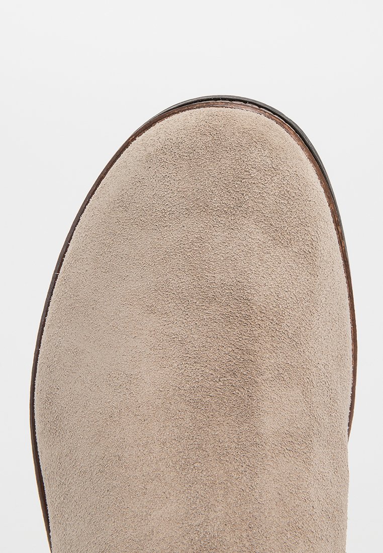 Beżowy zamszowy botek za kostkę z okrągłym noskiem, gładką fakturą i brązowym skórzanym wykończeniem wzdłuż podeszwy, widoczny z góry.