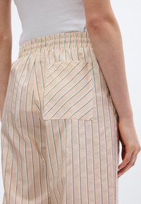 Ανοιχτά μπεζ σατέν παντελόνια με κάθετες λευκές και μαύρες ρίγες, με ελαστική μέση και μία πίσω τσέπη.