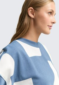 Jonge vrouw met lang blond haar in een blauw-witte gebreide trui, die naar rechts kijkt tegen een effen lichte achtergrond.