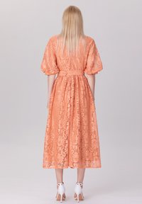 Robe midi en dentelle pastel orange avec des manches bouffantes, taille cintrée et jupe fluide. Elle présente une ceinture et des motifs floraux intricats.