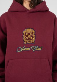 Bordowa bluza z kapturem z dużym haftowanym złotym i niebieskim herbem oraz napisem "Social Club" w stylizowanym kroju pisma. Posiada kieszeń z przodu i miękką teksturę.