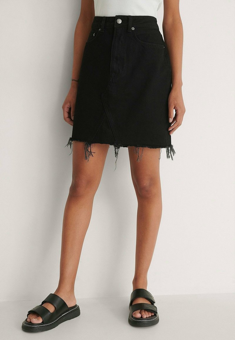 Femme portant une mini-jupe en denim noire avec un ourlet effiloché et des sandales slides noires, debout devant un mur clair uni.