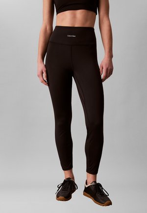 Frau trägt schwarze hoch taillierte Leggings von Calvin Klein und schwarze Sneakers mit hellbraunen Sohlen, steht vor einem grauen Hintergrund.