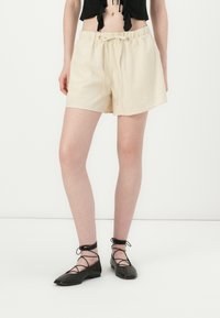 Beige bomullsshorts med elastisk midje og snøring. Modellen har på seg svarte flate sko med kryssende snøringer.