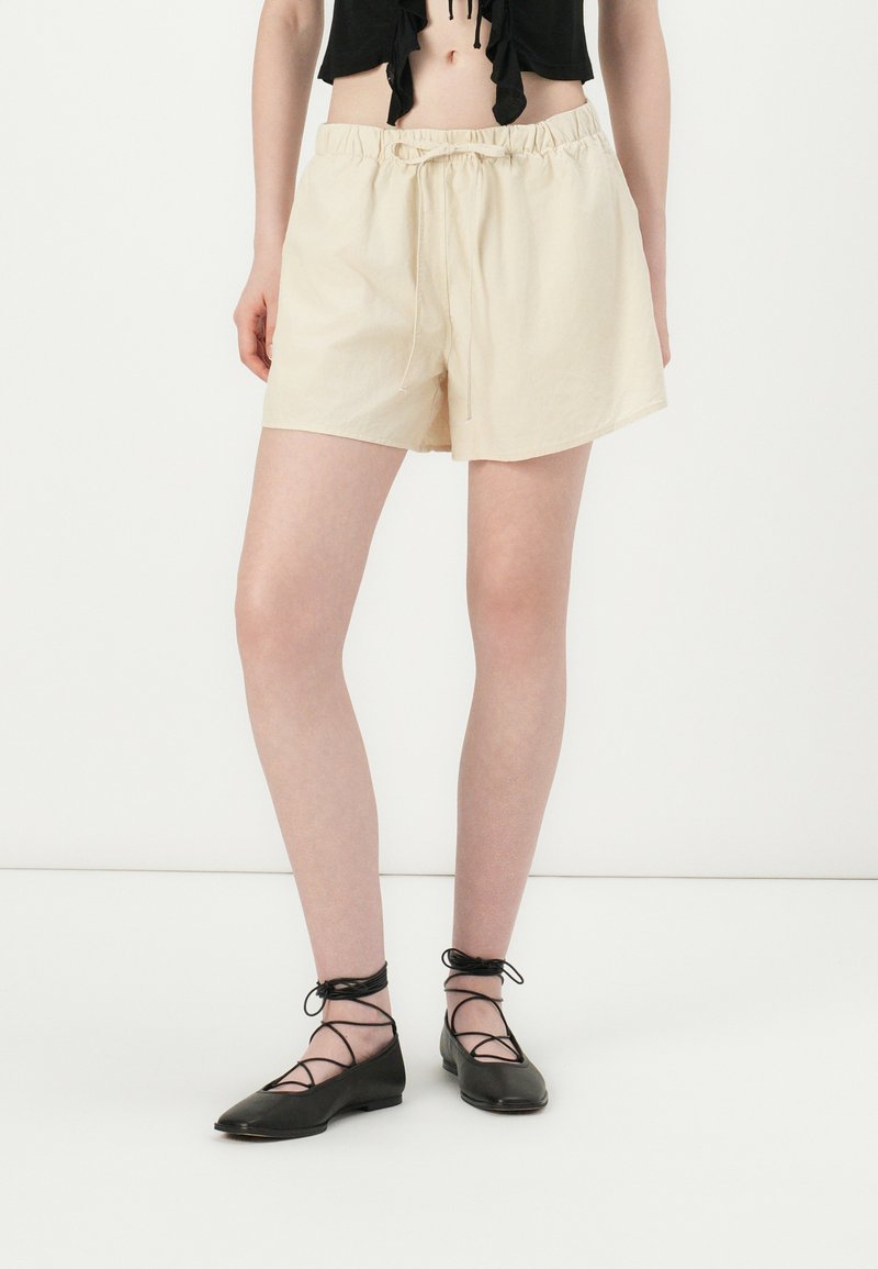 Beige bomullsshorts med elastisk midje og snøring. Modellen har på seg svarte flate sko med kryssende snøringer.