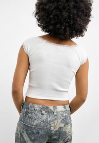 Hvit ribbestrikket cropped topp med blondekant ved halsen og korte ermer. Kombinert med teksturerte grå kamuflasjejeans med røde sømdetaljer.