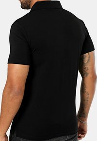 Camiseta polo negra con cuello, mangas cortas y una textura suave. Presenta una abertura lateral en el dobladillo y elementos de diseño mínimos.