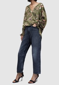 Blouse verte à motifs floraux avec de longues manches bouffantes, associée à un jean en denim foncé taille haute et coupe droite, ainsi qu'à des talons ouverts noirs.