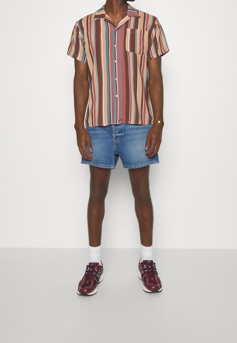 Homme portant une chemise à manches courtes à rayures verticales, un short en denim bleu, des chaussettes blanches et des baskets bordeaux, debout devant un fond blanc.