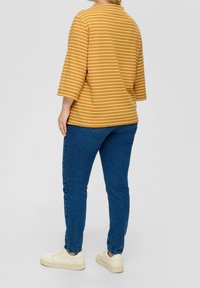 Gestreept geel shirt met driekwart mouwen, gecombineerd met blauwe spijkerbroek. Model draagt witte sneakers, wat een ontspannen, casual outfit laat zien.