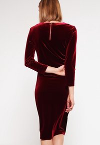 Robe en velours bourgogne à manches longues, avec une coupe ajustée et une fermeture éclair à l'arrière. Texture lisse et design jusqu'aux genoux.