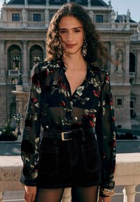 Transparente schwarze Bluse mit rotem Rosenmuster, kombiniert mit hoch geschnittenen schwarzen Shorts und einem schwarzen Gürtel. Verfügt über lange Ärmel und dezente Hardware-Akzente.