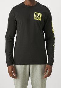 T-shirt a maniche lunghe nera in cotone, con una grafica gialla "NIKE HOOPS" sul petto e lettering giallo lungo la manica sinistra.