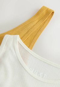 Gros plan sur un débardeur blanc côtelé superposé à un débardeur moutarde côtelé sur fond clair.