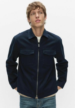 Ung man klädd i marinblå jacka med dragkedja och bröstfickor samt randig tröja under, tittar åt sidan med händerna i fickorna.