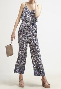 Kobieta w bezrękawowym kwiatowym kombinezonie z szerokimi nogawkami, trzymająca beżową kopertówkę i nosząca beżowe platformy.