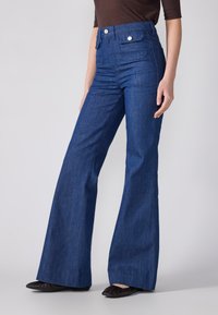 Τζιν σε στυλ καμπάνα σε σκούρο μπλε, με ψηλή μέση και δύο πίσω τσέπες με ασημί υλικό. Λείο ύφασμα και κλασικός σχεδιασμός.