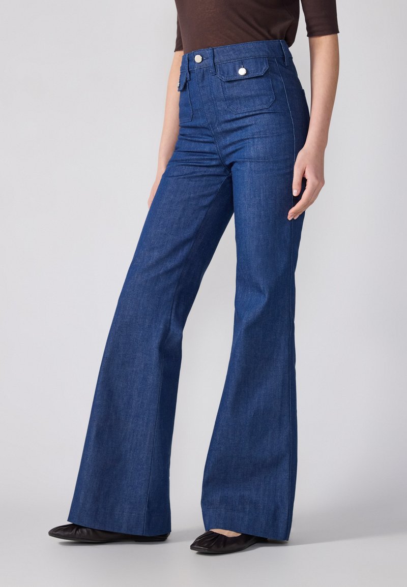 Τζιν σε στυλ καμπάνα σε σκούρο μπλε, με ψηλή μέση και δύο πίσω τσέπες με ασημί υλικό. Λείο ύφασμα και κλασικός σχεδιασμός.