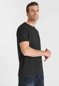 Uomo che indossa una semplice t-shirt nera a maniche corte e jeans scuri, che guarda di lato con le mani unite davanti a uno sfondo bianco.