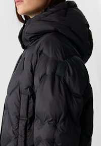 Kobieta ubrana w czarną pikowaną kurtkę puchową z kapturem, z delikatnym ukośnym przeszyciem i małą naszywką na górnej części rękawa.