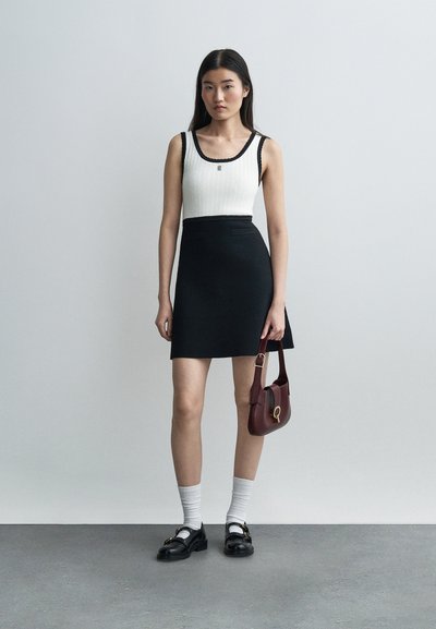 Γυναίκα στέκεται μπροστά σε γκρίζο τοίχο, φορώντας λευκή αμάνικη μπλούζα, μαύρη φούστα, λευκές κάλτσες, μαύρες μοκασίνες και κρατώντας μια σκούρα κόκκινη τσάντα χειρός.