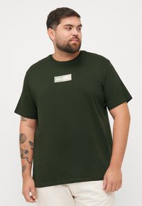 Tamnozeleni pamučni majica s kratkim rukavima, okruglim izrezom i "JACK & JONES" logotipom u pravokutnom jastučiću sprijeda. Glatka tekstura.